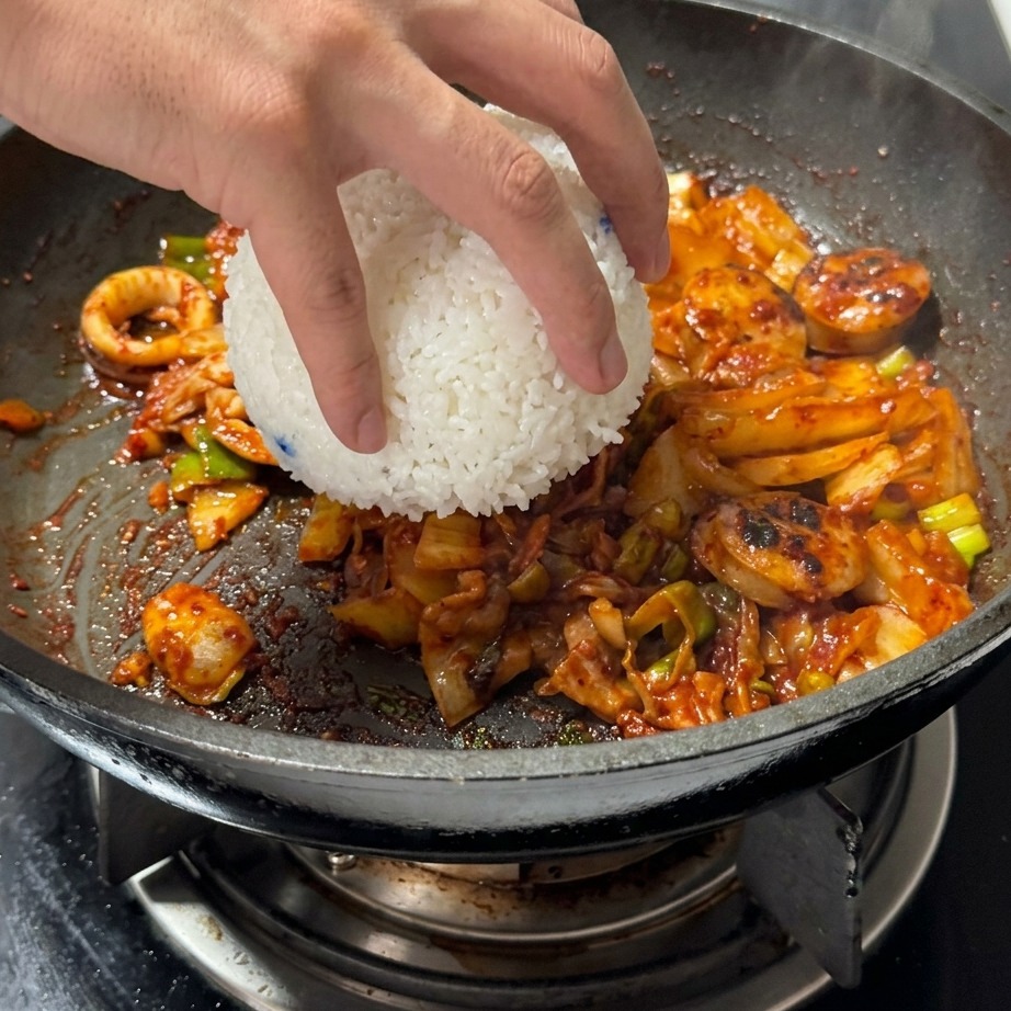 Una ciotola di riso bianco cotto al vapore che viene aggiunta a una padella contenente kimchi e carne saltati.