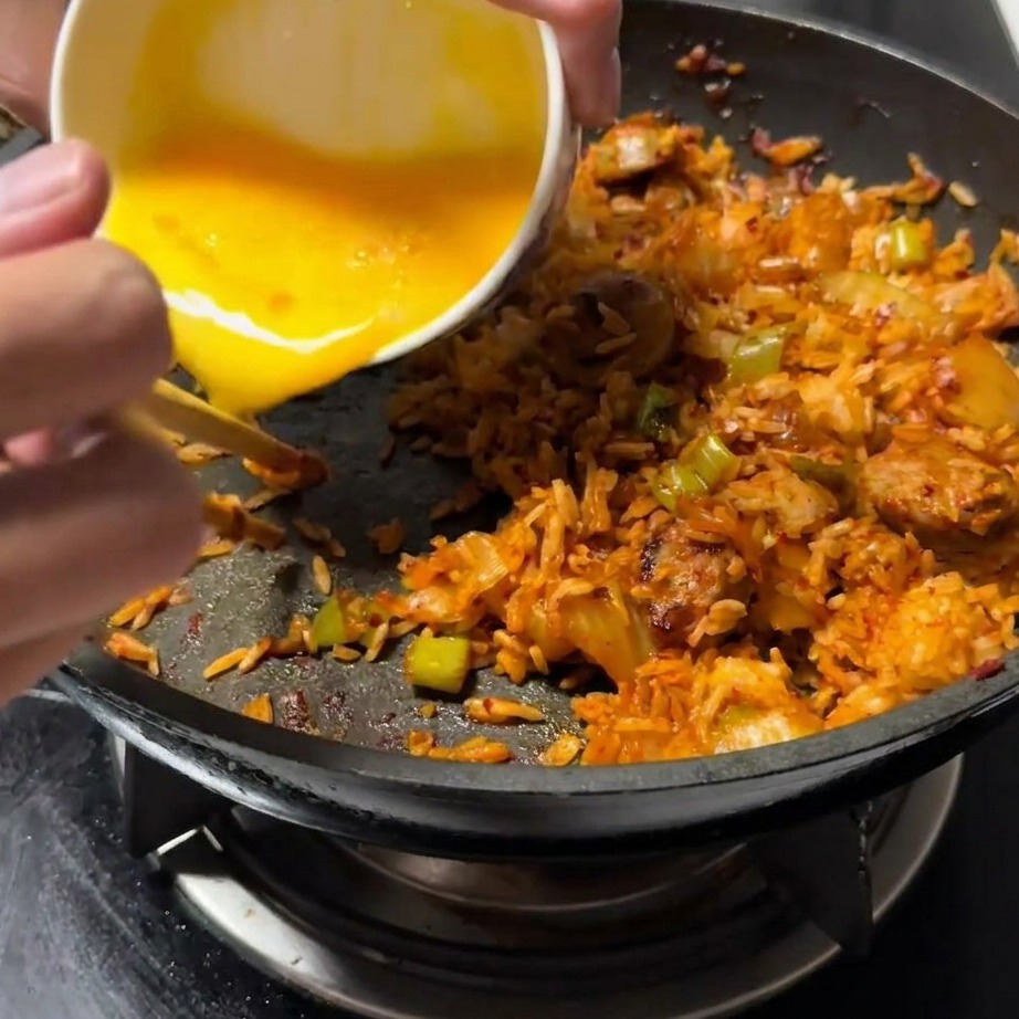 Uovo sbattuto che viene versato in un lato libero della padella, accanto al composto di riso fritto.