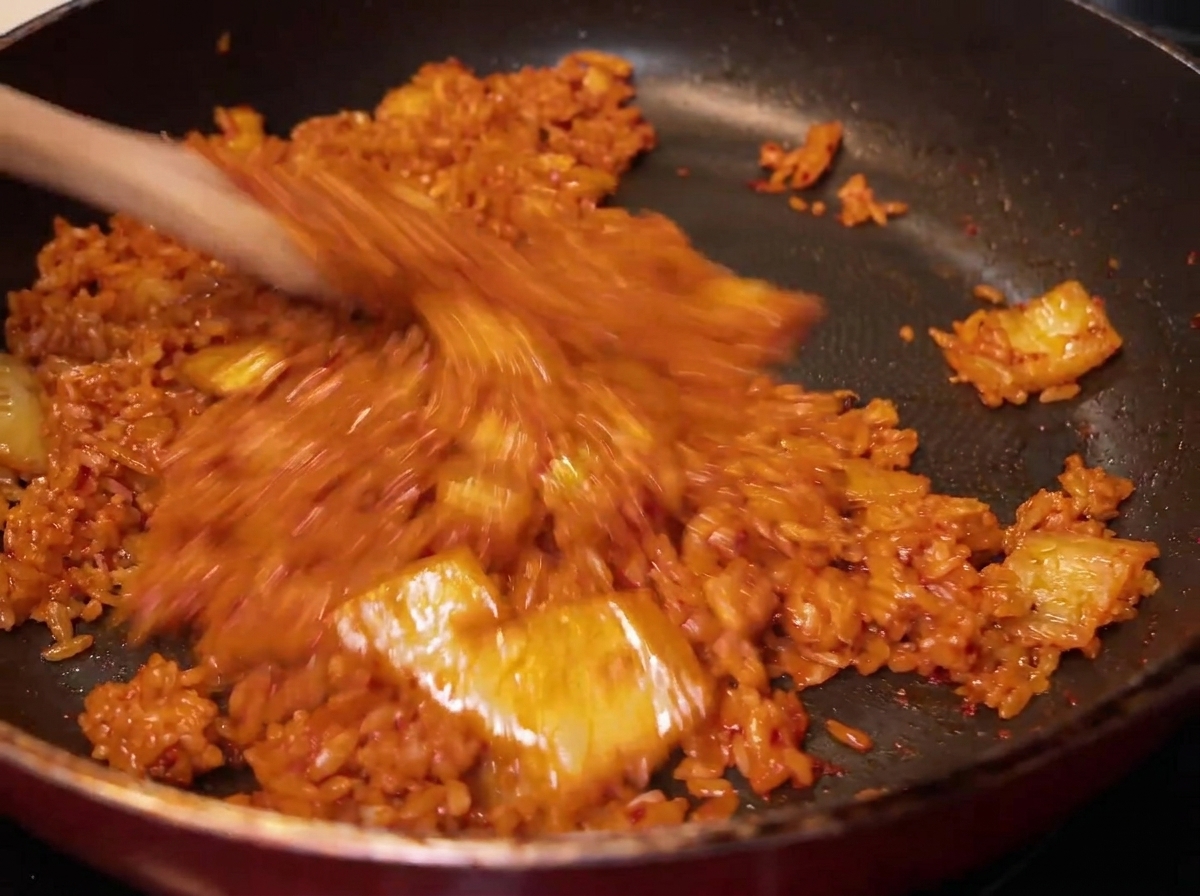 Primo piano del riso mescolato con il kimchi nella padella, che assume un colore rosso-arancio uniforme.