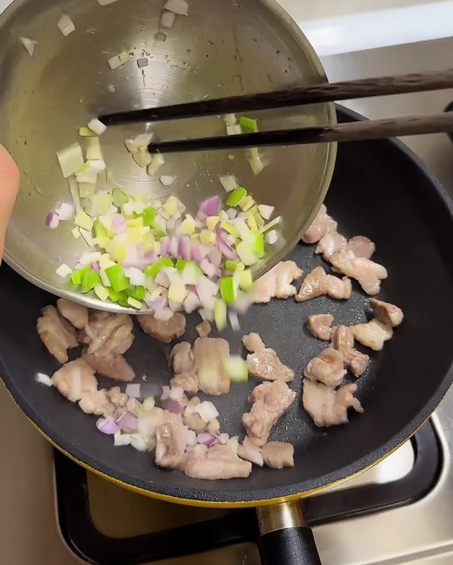 Cipollotti, aglio e cipolla tritati versati da una ciotola nella padella con la pancetta cotta.