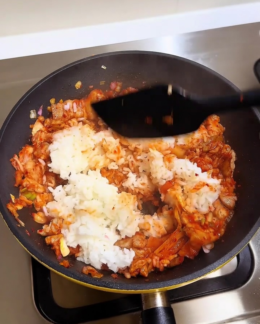 Una spatola che mescola riso bianco in una base vibrante rossa di maiale e kimchi in una padella calda.