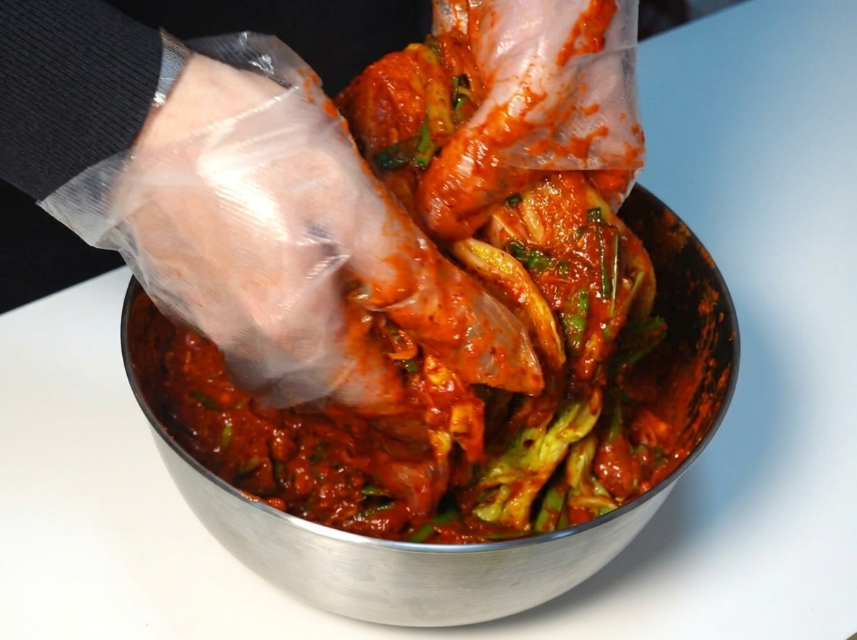 Mani guantate che spalmano una densa pasta di kimchi rossa uniformemente su ogni strato di una testa di cavolo napa.
