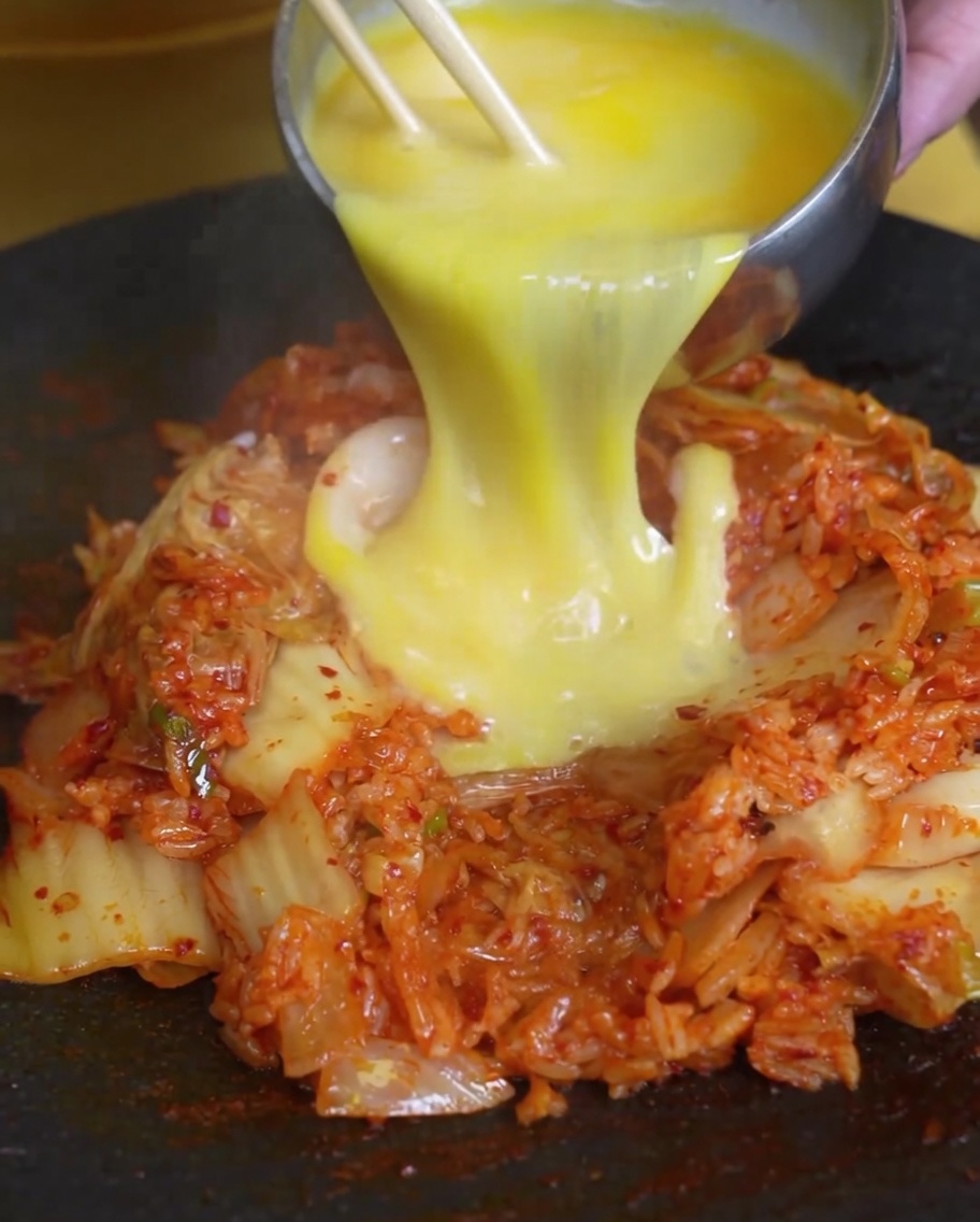 Uova sbattute versate sopra un mucchietto di riso saltato al kimchi in una padella calda.