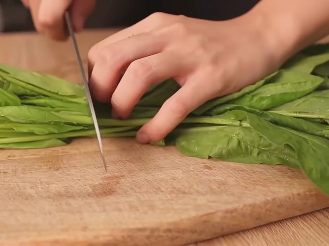 Una mano che taglia un mazzetto di spinaci freschi in sezioni su un tagliere di legno.