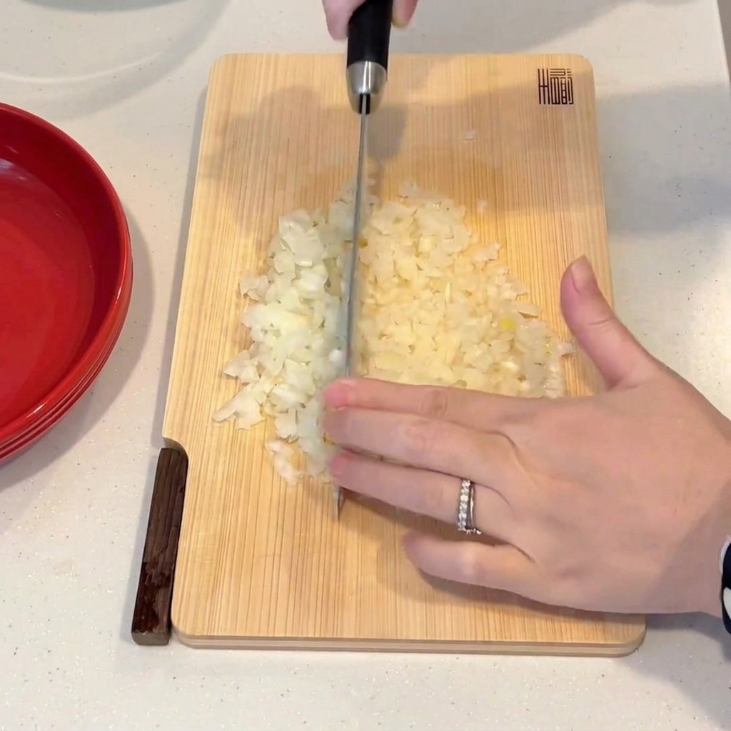 Una mano che usa un coltello da chef per tritare finemente una cipolla su un tagliere di legno.