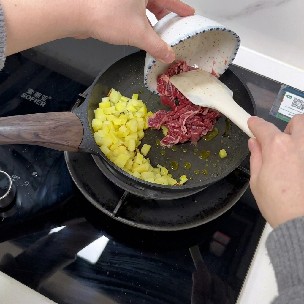 Fettine di manzo aggiunte in una padella con patate cotte usando una spatola bianca.