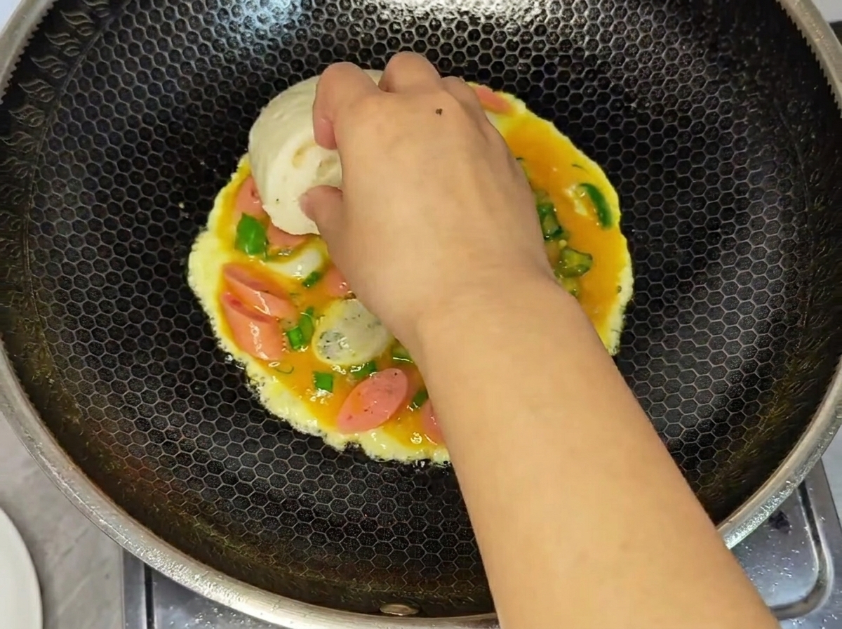 Una mano preme mezza porzione di panino al vapore sulle uova ancora crude nel wok.