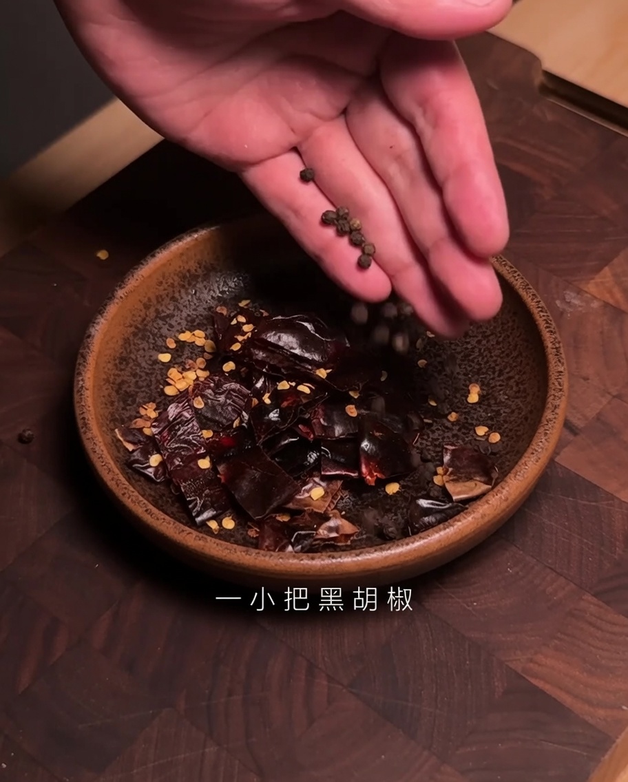 Mani che versano grani di pepe nero in una ciotola contenente pezzi di peperoncino Guajillo tagliato.