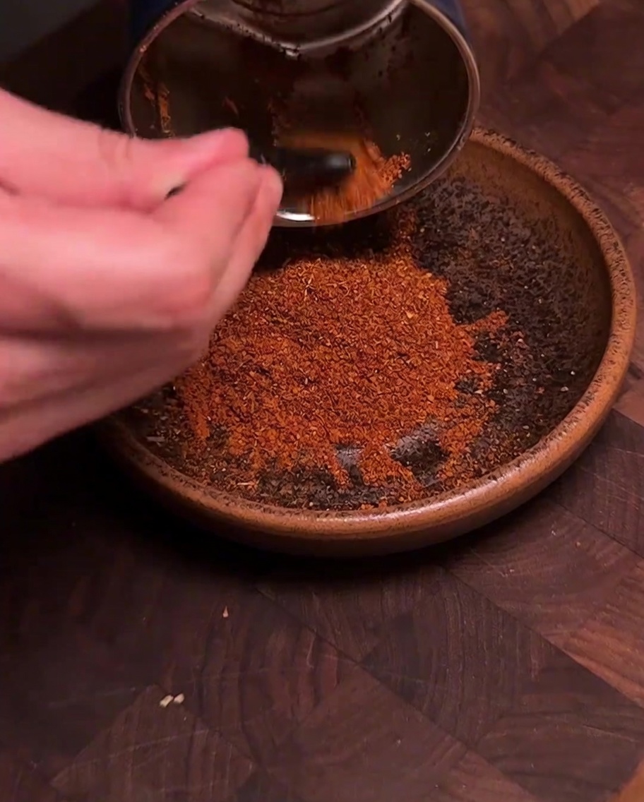 Un macinaspezie che versa polvere di peperoncino e spezie di colore rosso vivo appena macinata in una ciotola di ceramica marrone.