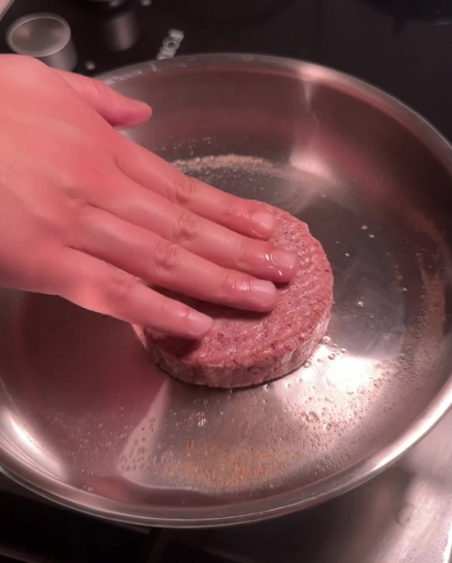 Un hamburger di manzo crudo che viene premuto in una padella calda in acciaio inox per la scottatura.