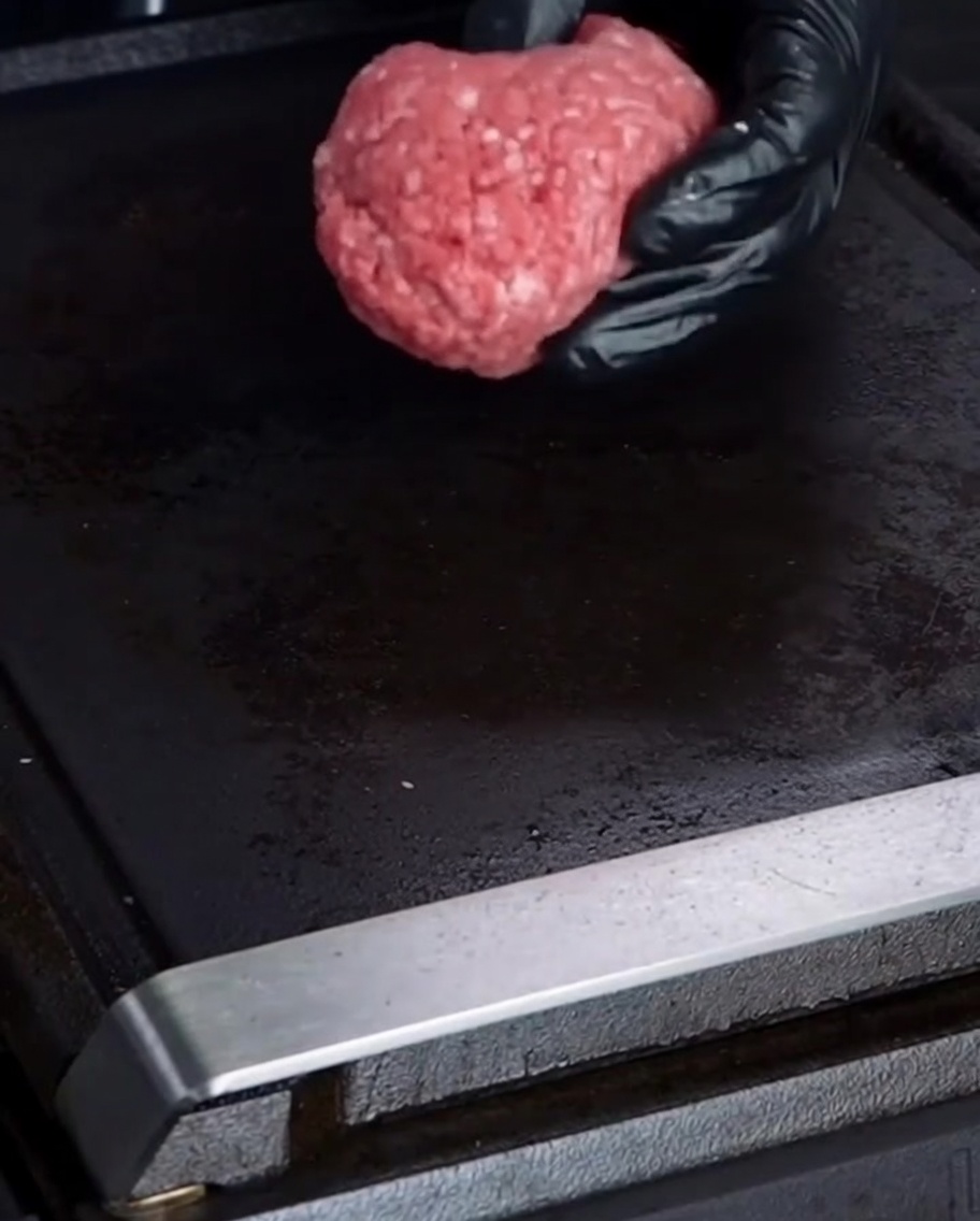 Una mano guantata che posiziona una pallina di carne di manzo 75/25 cruda su una piastra nera.