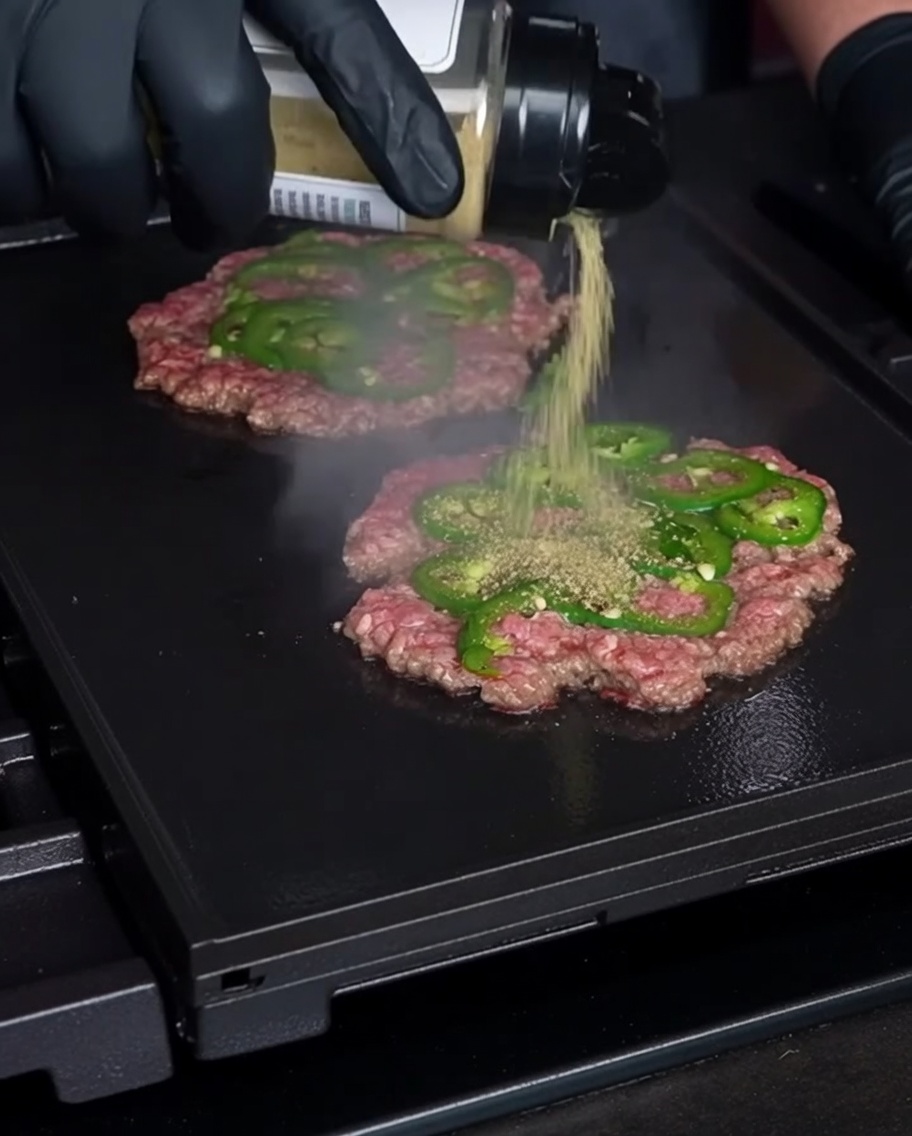 Una mano guantata che sparge del condimento su sottili burger di manzo con fettine di jalapeño su una piastra calda.