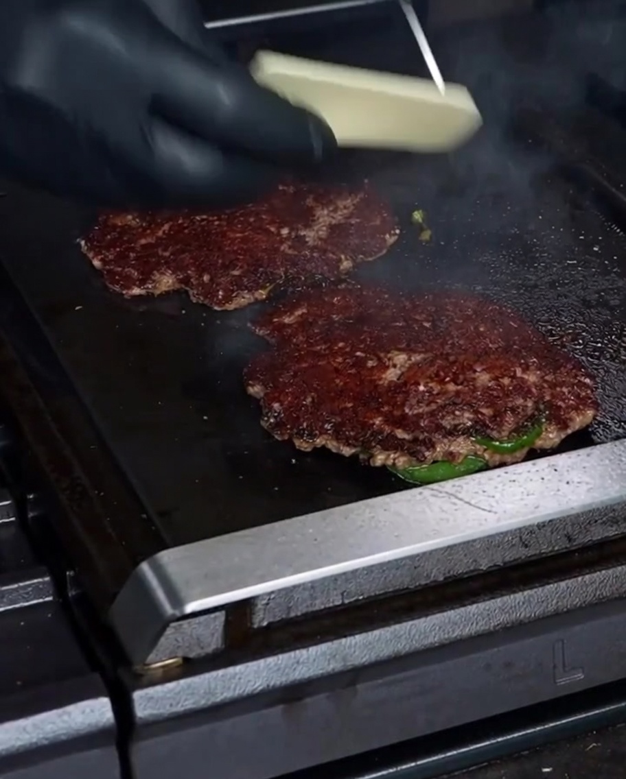 Una mano guantata che posiziona una fetta di formaggio Havarti su un burger ben rosolato sulla piastra.