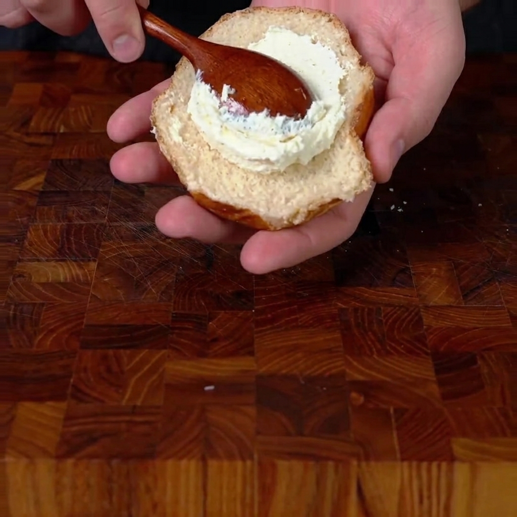 Un cucchiaio di legno che spalma un generoso strato di formaggio spalmabile montato sulla base di un panino tostato.