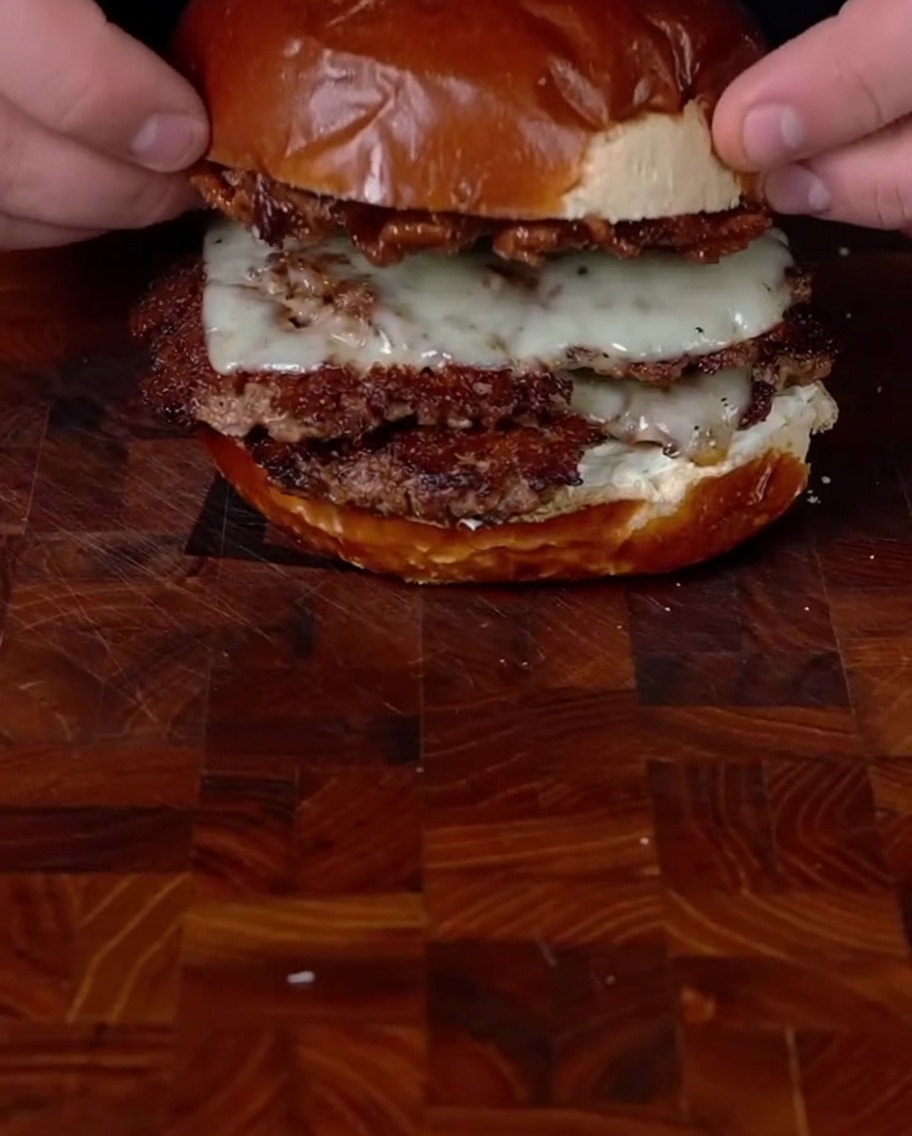 Mani che posizionano la parte superiore del panino con la confettura sul doppio smashburger per completare lassemblaggio.