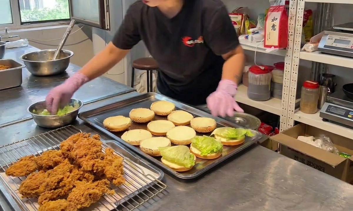 Una persona posiziona un pezzo di pollo fritto croccante sulla base di un panino per hamburger ricoperta di lattuga verde.