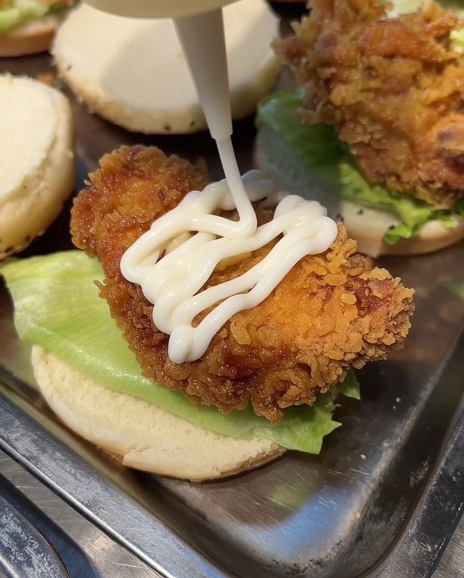 Maionese bianca spremuta da un dosatore su un pezzo di pollo fritto adagiato su un panino da burger.