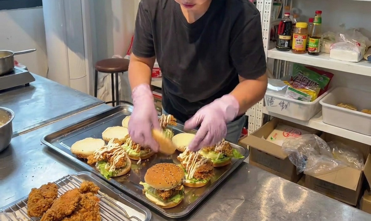 Una persona posiziona la metà superiore di un panino al sesamo su un burger di pollo fritto completamente assemblato.