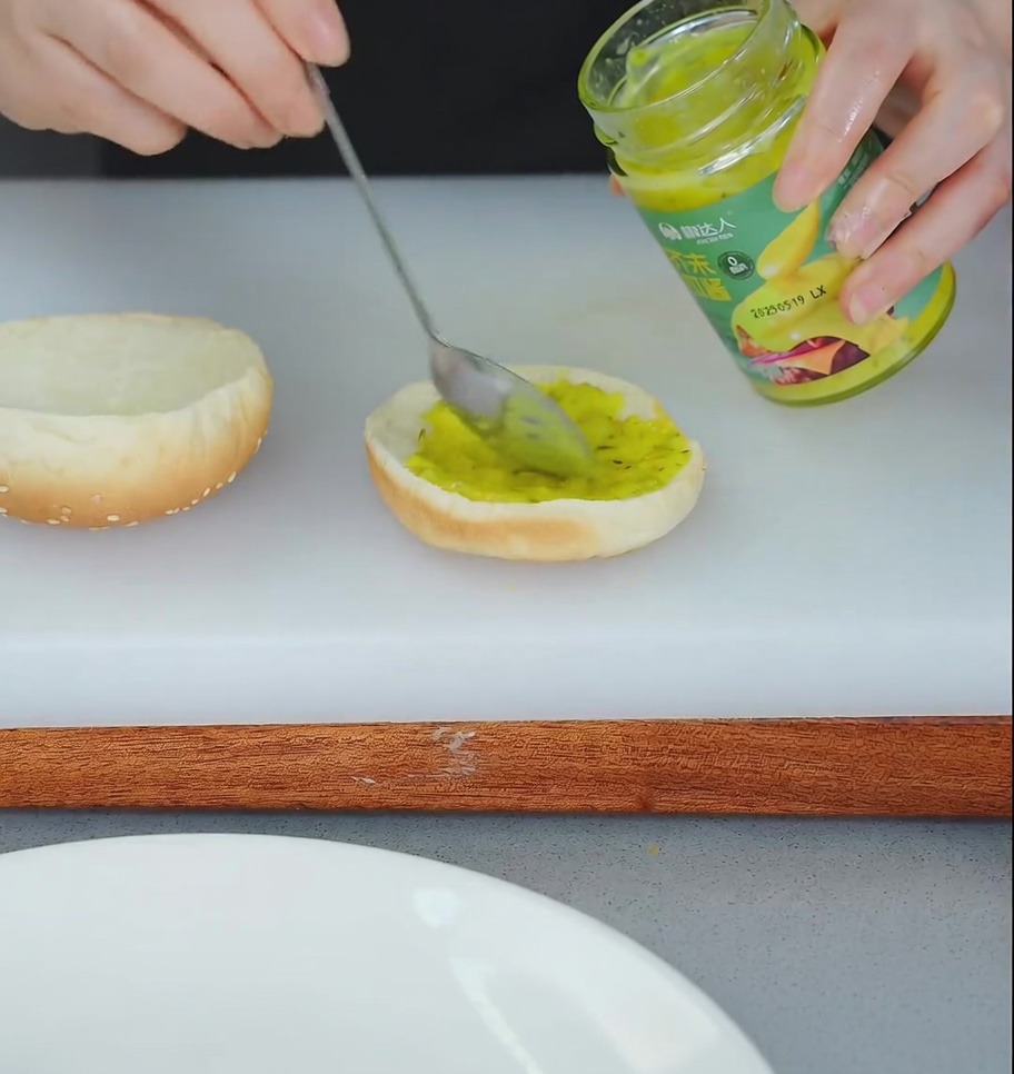 Una persona spalma del relish giallo sulla base di un panino per hamburger tostato.
