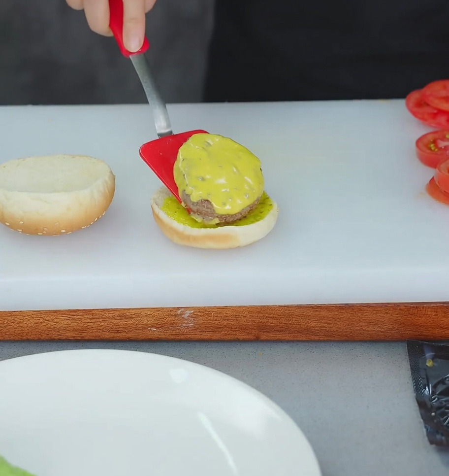 Una spatola rossa che trasferisce lhamburger col formaggio sul pane tostato già guarnito.