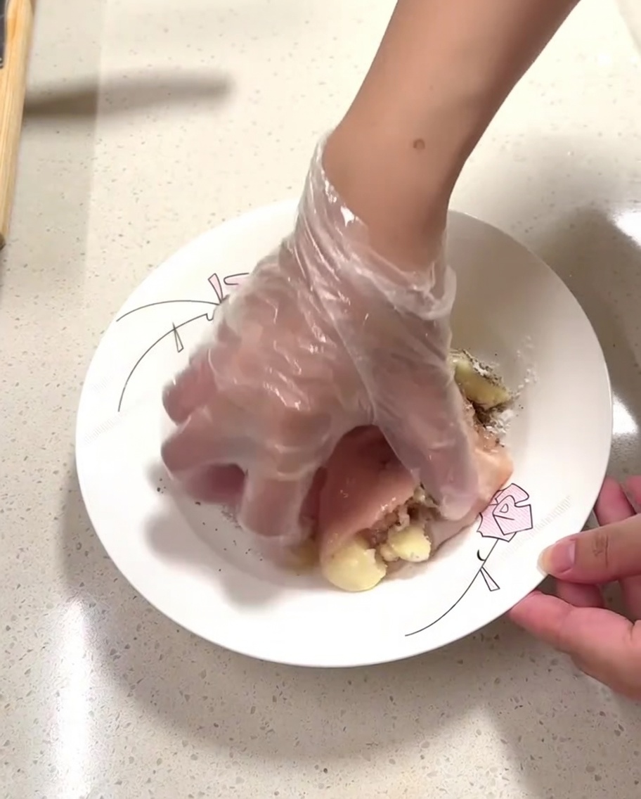 Una mano con un guanto massaggia i condimenti sui pezzi di pollo crudo in un piatto.