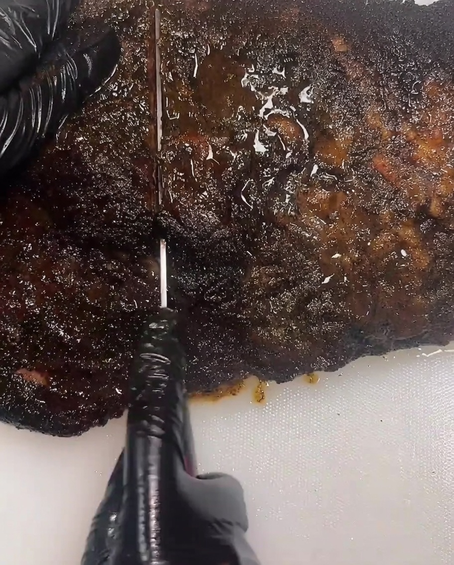 Primo piano di una persona con guanti neri che usa un lungo coltello per affettare il brisket su un tagliere bianco.