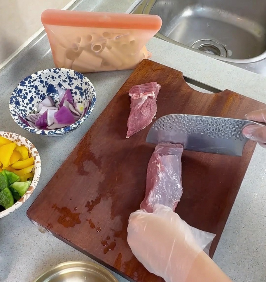 Mani con guanti che usano una mannaia per tagliare il maiale crudo a striscioline su un tagliere di legno.