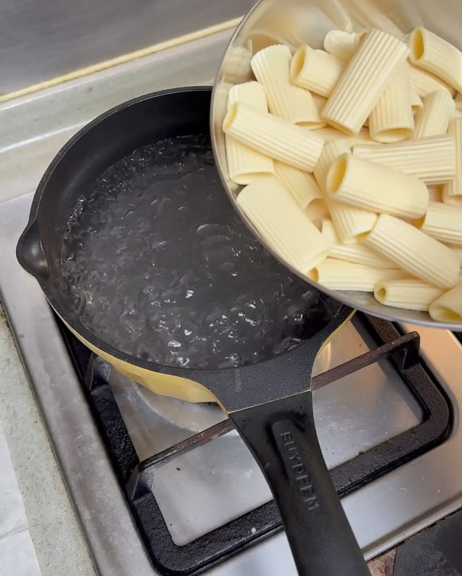 Rigatoni precedentemente ammollati versati da una ciotola di metallo in una pentola di acqua bollente sui fornelli.