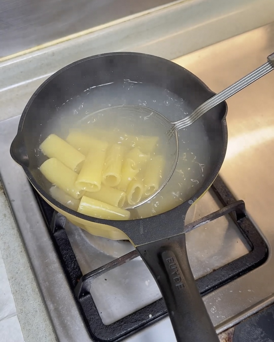 Una schiumarola a rete che preleva i rigatoni cotti dallacqua bollente in una pentola nera.