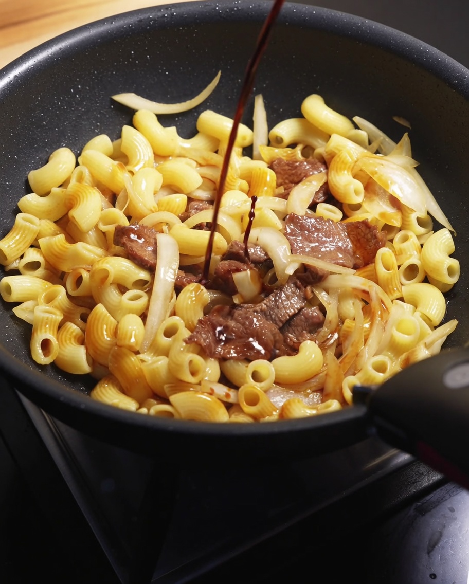 Un flusso di salsa di soia scura versato sopra il composto di manzo e maccheroni in una padella calda.