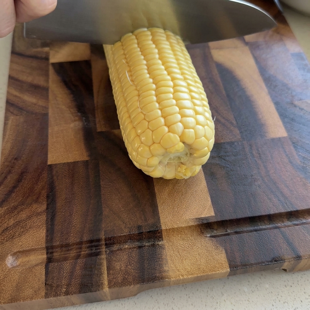 Una mano che usa un grande coltello da chef per affettare chicchi di mais giallo crudo da una pannocchia su un tagliere di legno.