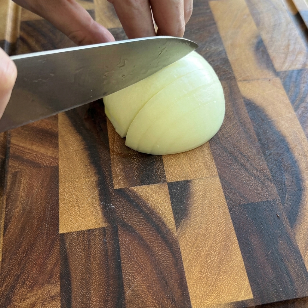 Una persona che affetta mezza cipolla bianca in piccoli pezzi su un tagliere di legno.