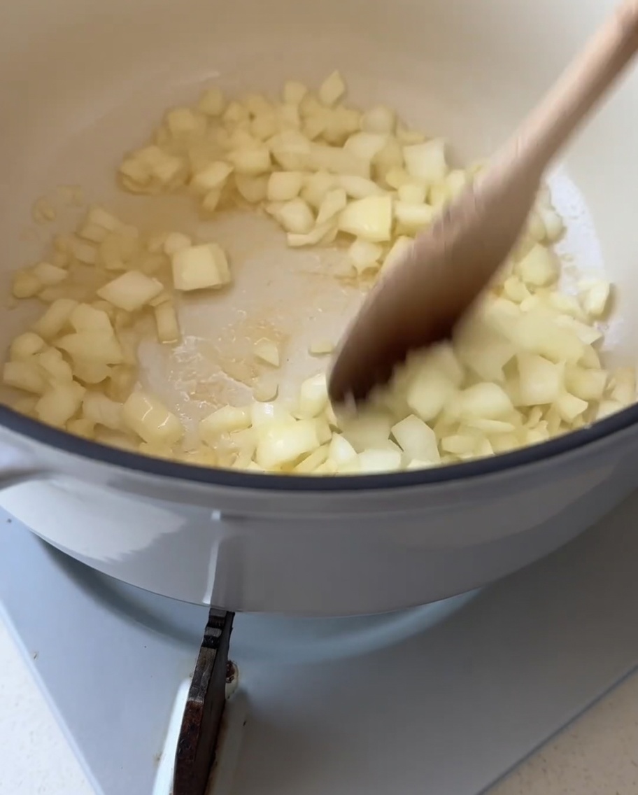 Un cucchiaio di legno che mescola cipolle bianche tritate allinterno di una pentola bianca con burro fuso.