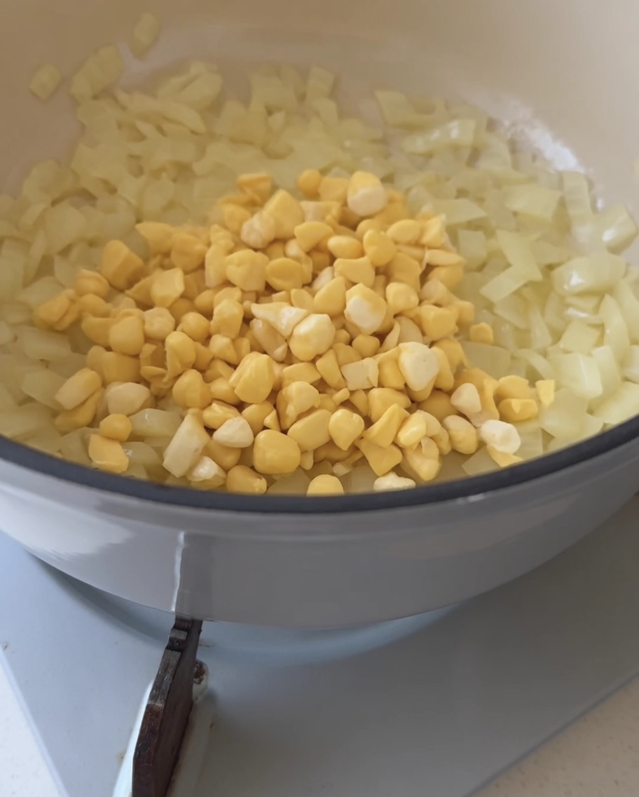 Chicchi di mais giallo fresco versati in una pentola contenente cipolle soffritte traslucide.