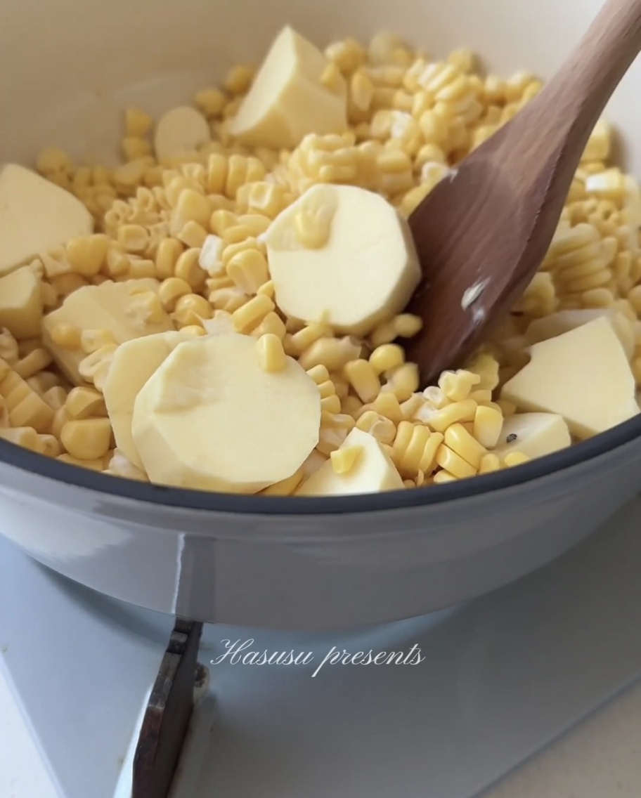 Un cucchiaio di legno che mescola un mix di chicchi di mais giallo e cubetti di patata dolce bianca in una pentola.