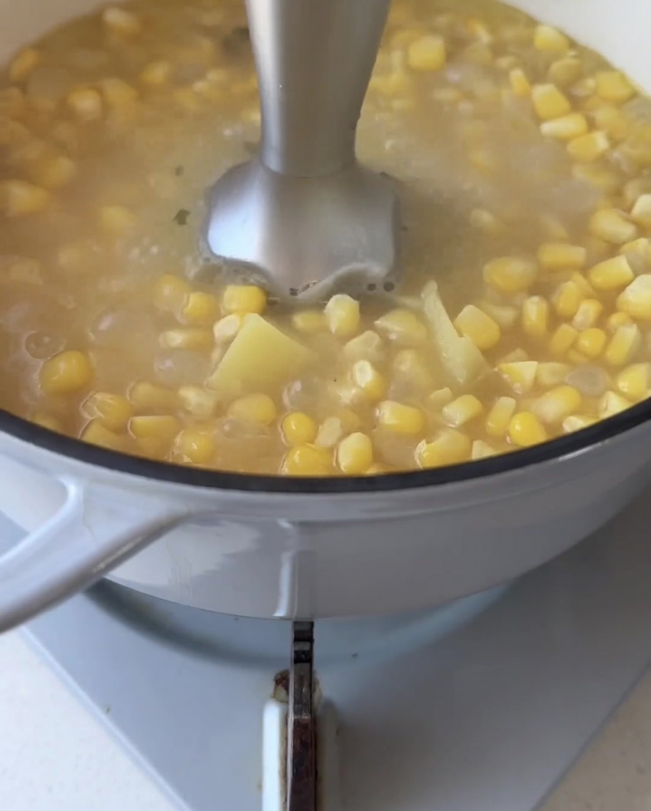 Un frullatore a immersione immerso in una pentola di chicchi di mais e cubetti di patata dolce che sobbollono, frullandoli in un liquido liscio.