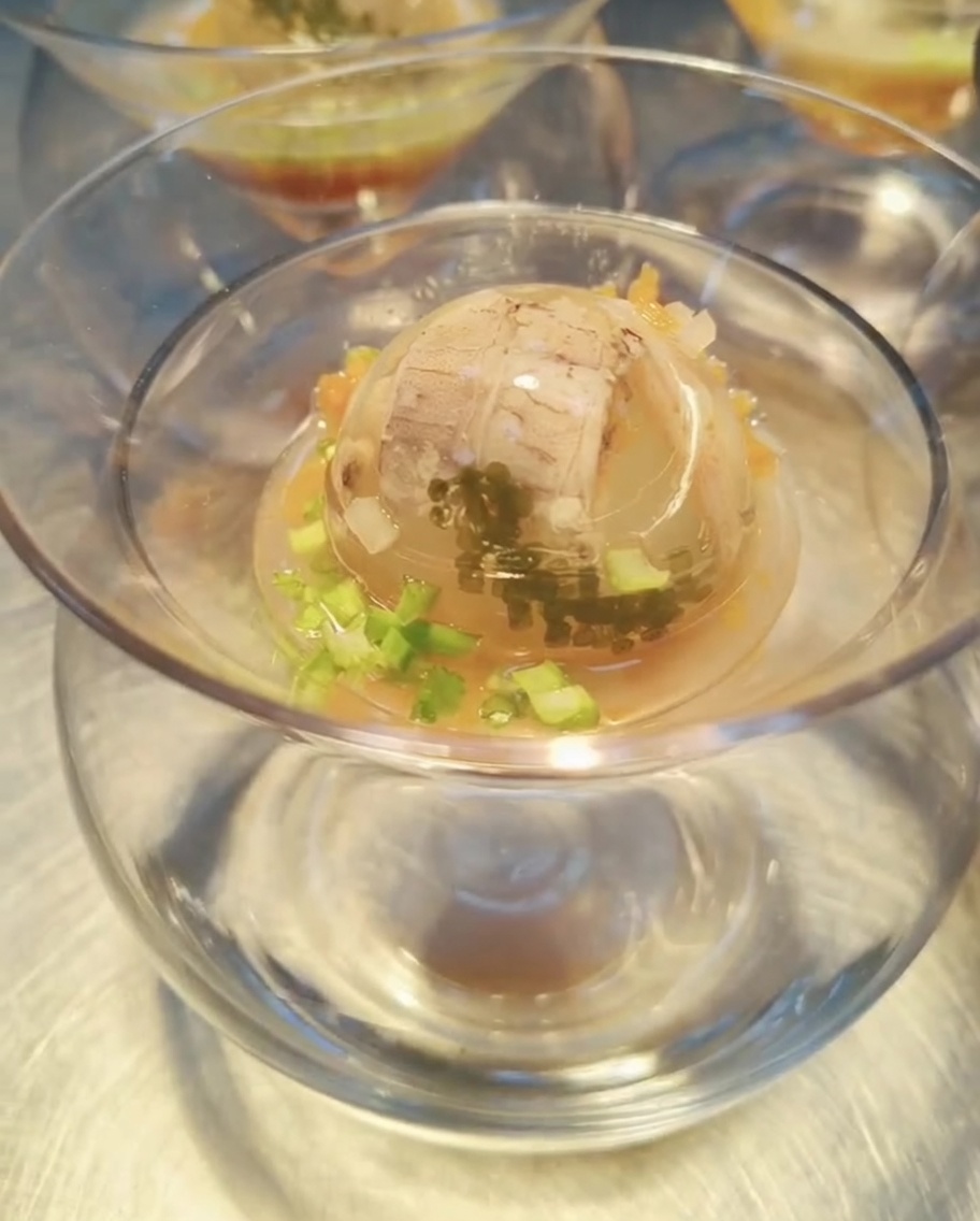 Una sfera di gelatina ai frutti di mare perfettamente rappresa in una ciotola di vetro, su cui viene versata una ricca salsa ambrata, pronta per essere guarnita e servita.