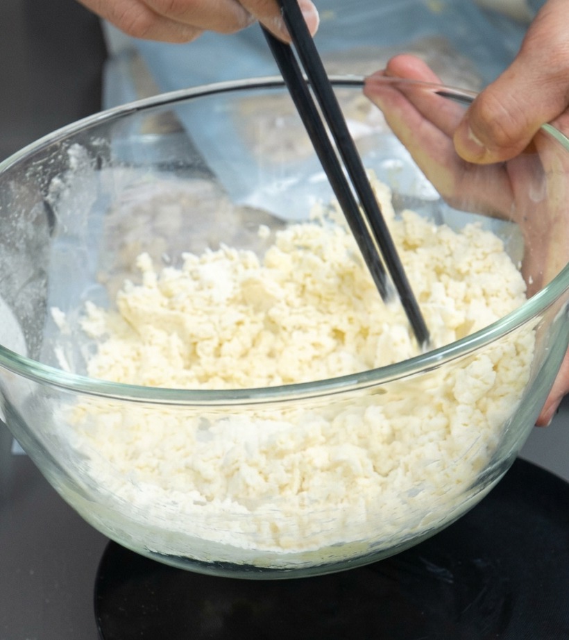 Una ciotola contenente un composto di farina e uova mescolato con le bacchette per formare delle briciole.