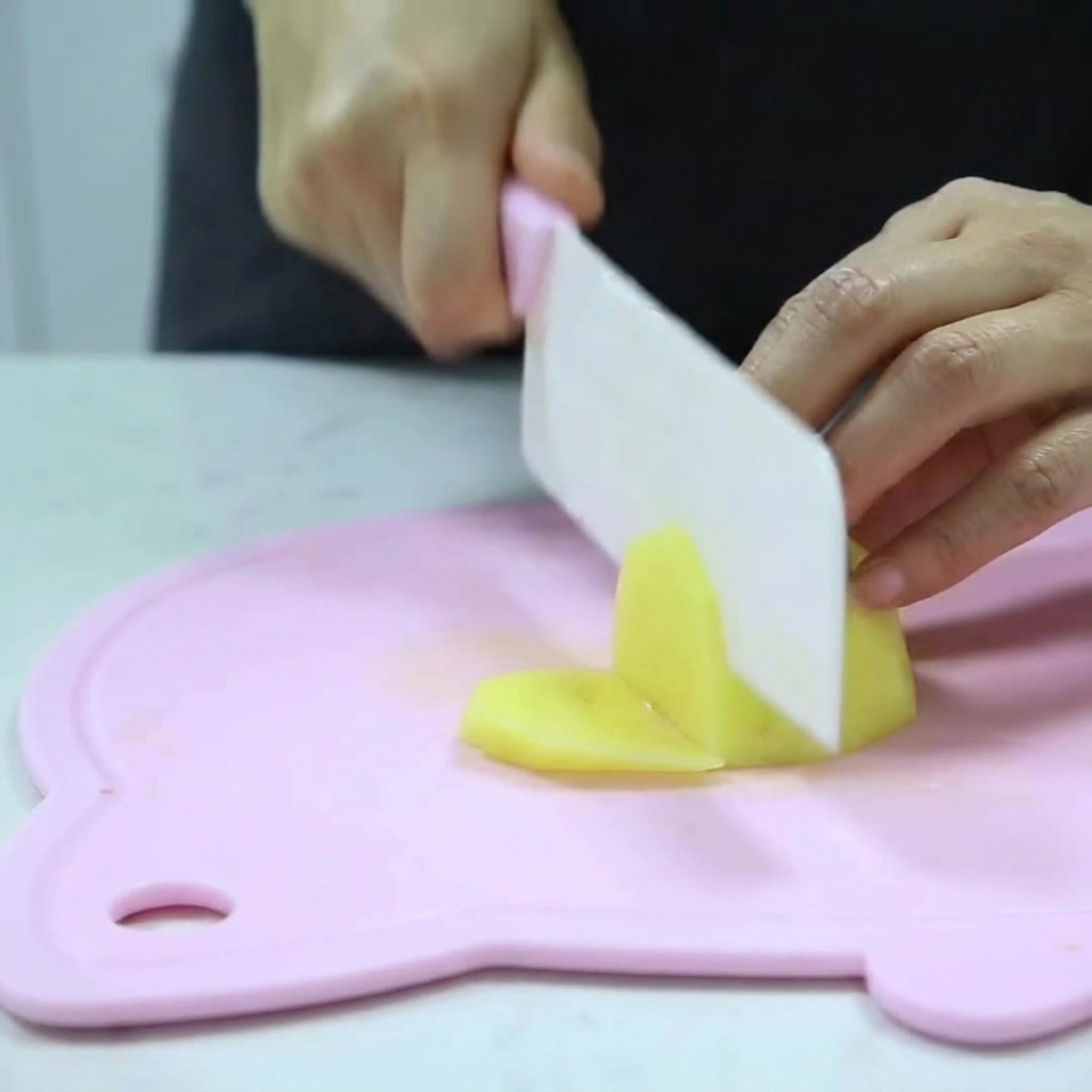 Mani che impugnano un coltello per tagliare una patata sbucciata a piccoli cubetti su un tagliere rosa.