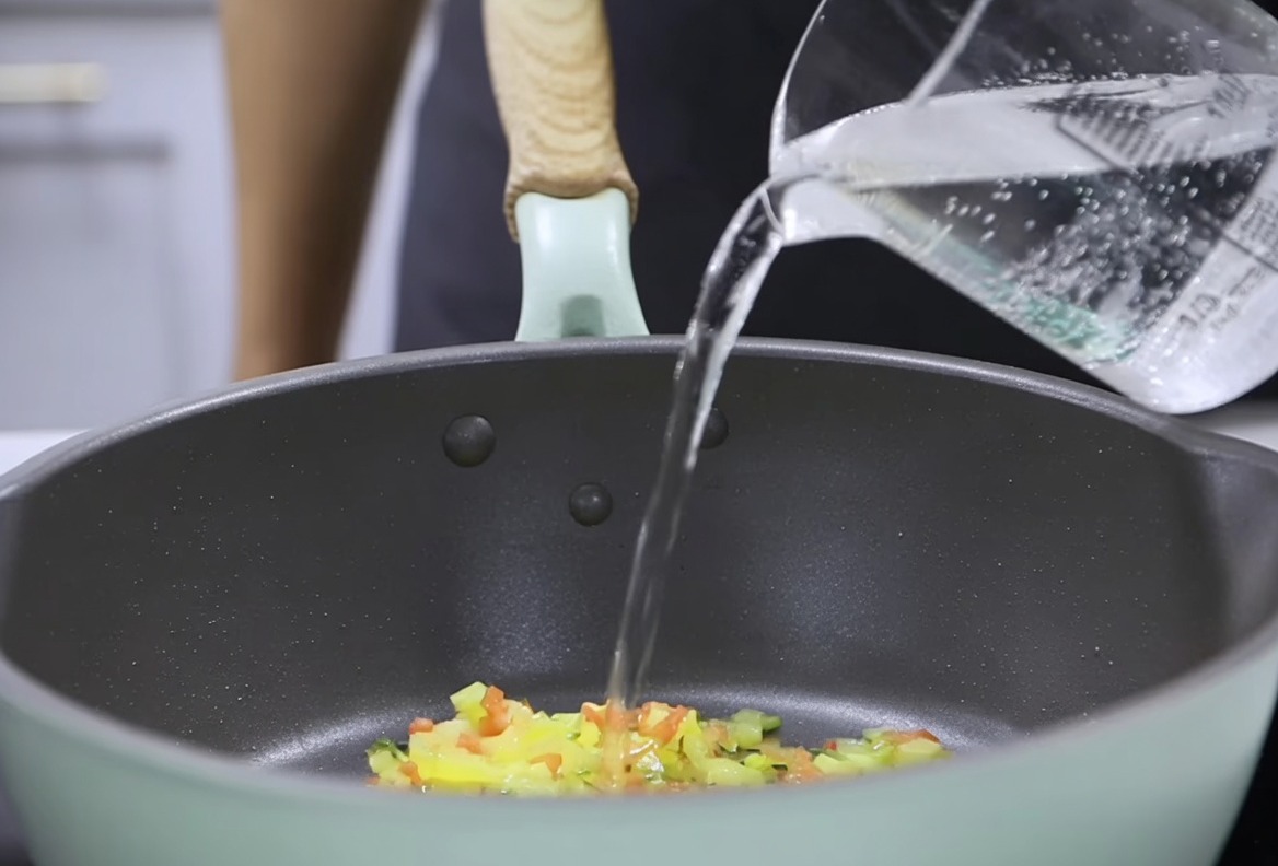 Acqua versata da un misurino in una padella con le verdure saltate, pronta per incorporare la crema di latte.