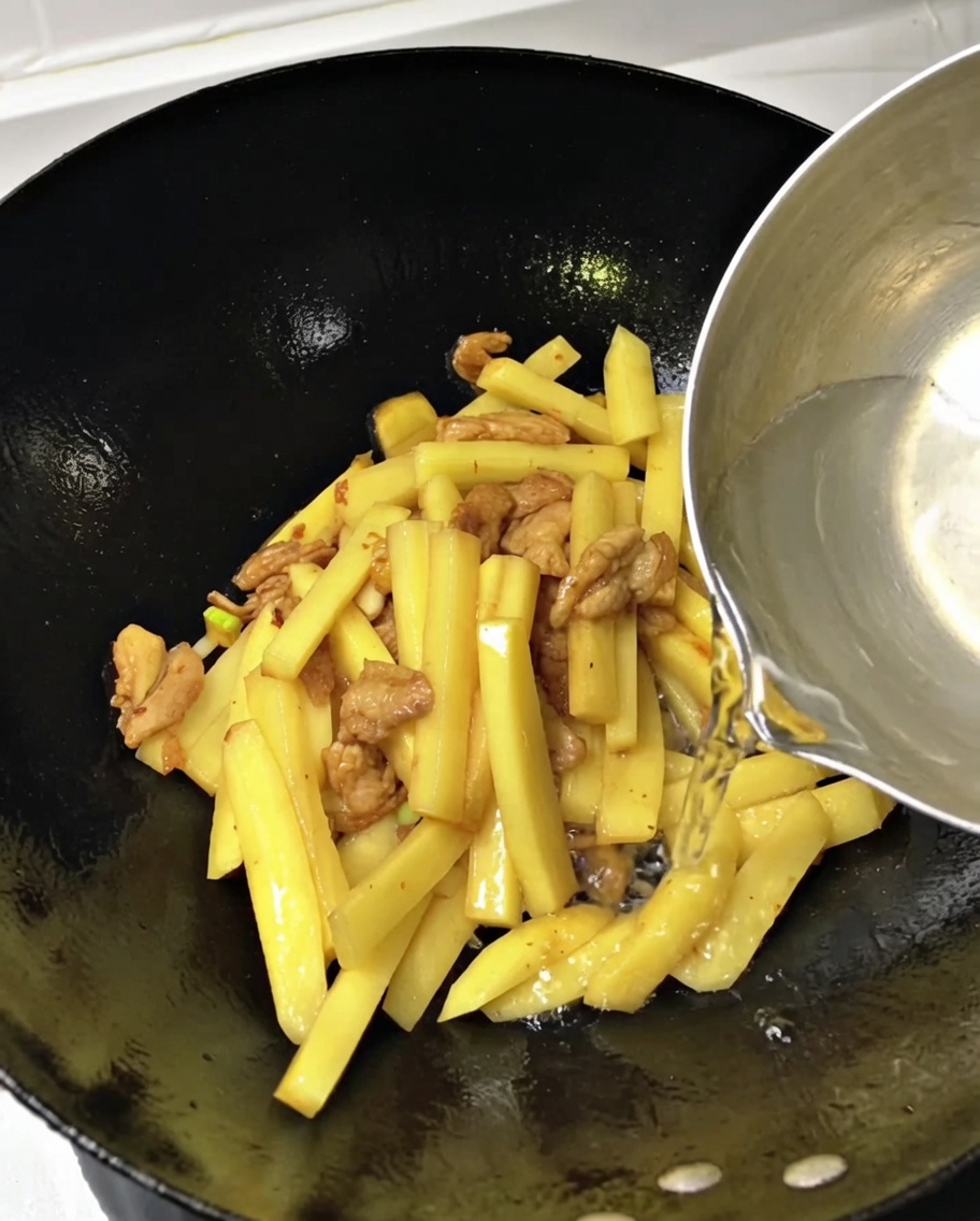 Acqua versata da un mestolo in un wok pieno di patate a fiammifero e carne di maiale.