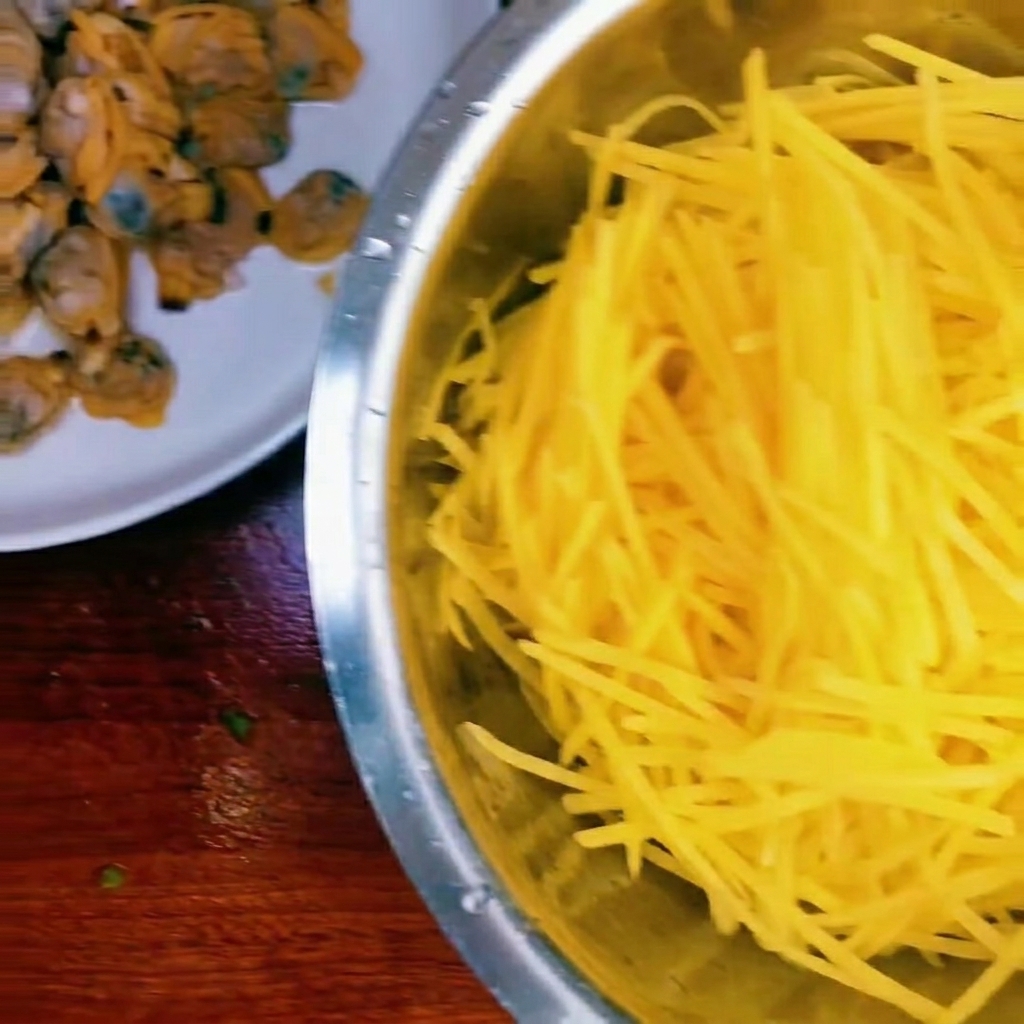 Una ciotola contenente patate crude a filetti, polpa di vongole cotta e un piccolo mucchio di cipollotti tritati.