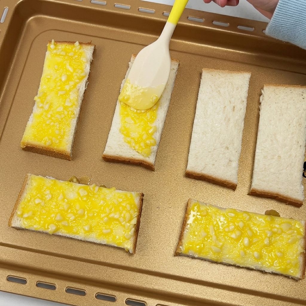 Una spatola che spalma burro allaglio su fette di pane posizionate su una teglia.