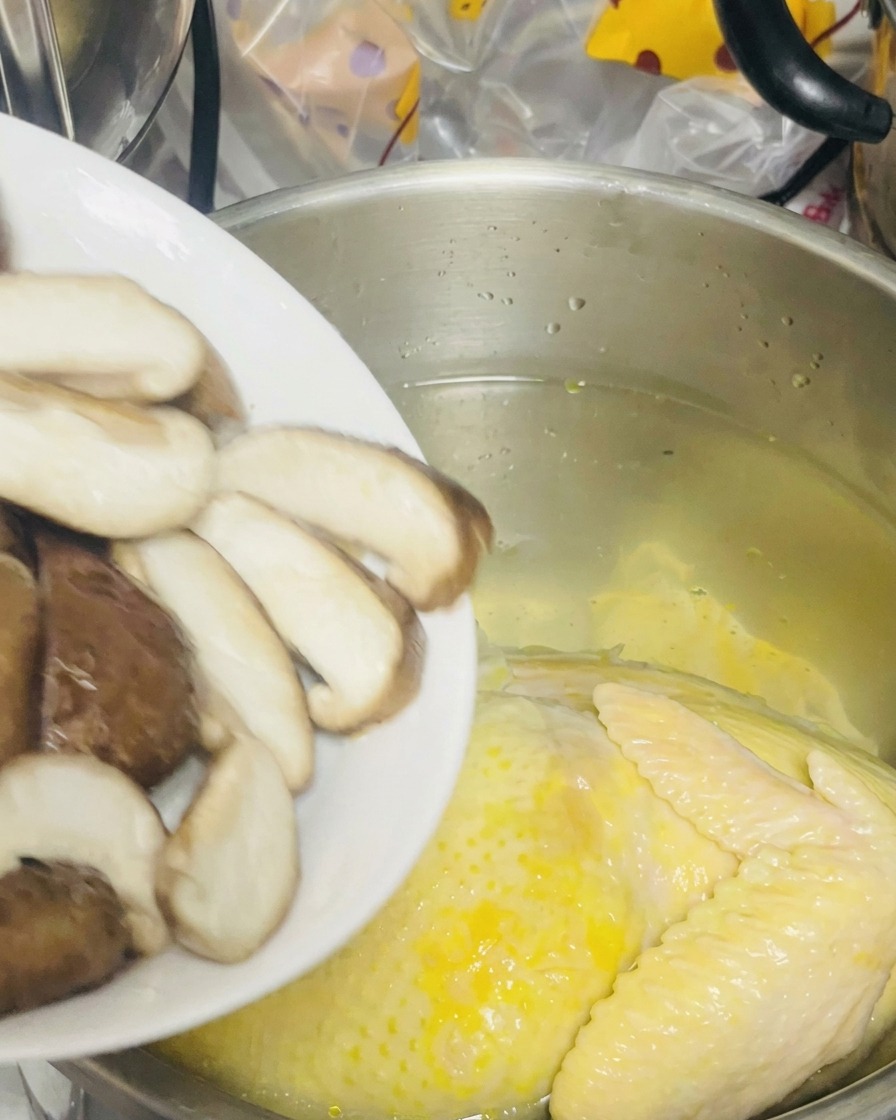 Funghi shiitake affettati versati da un piatto in una pentola contenente il pollo intero in acqua.