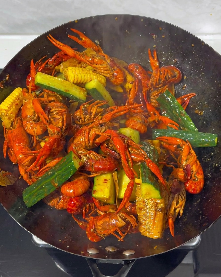 Un primo piano di gamberi, mais e pezzi di cetriolo mescolati insieme in un grande wok con una salsa rosso scuro ricca.