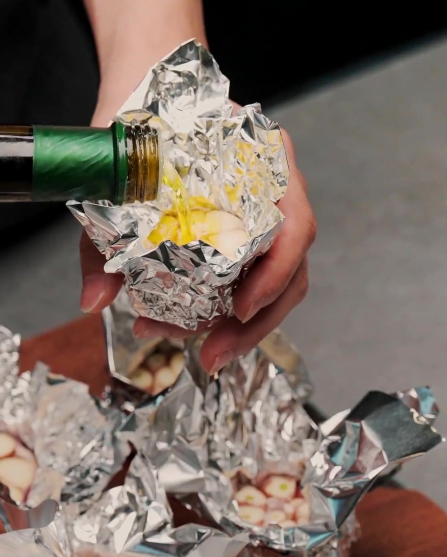 Olio doliva versato da una bottiglia verde sopra gli spicchi daglio esposti su un foglio di carta stagnola.