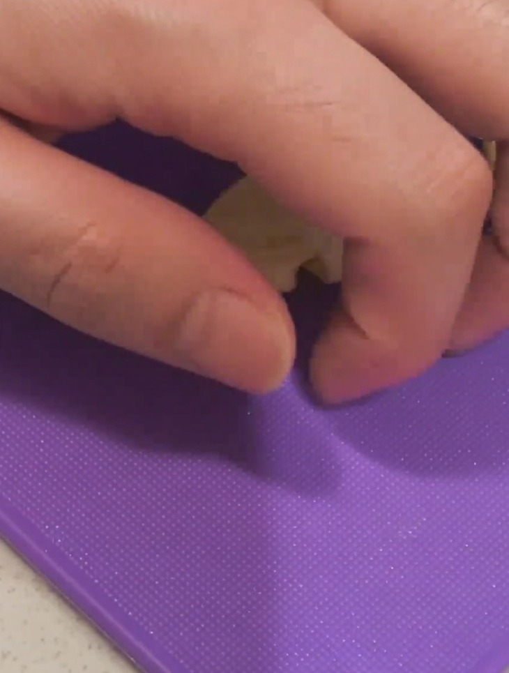 Primo piano di una mano che schiaccia uno spicchio daglio su un tagliere viola usando la lama di un coltello.