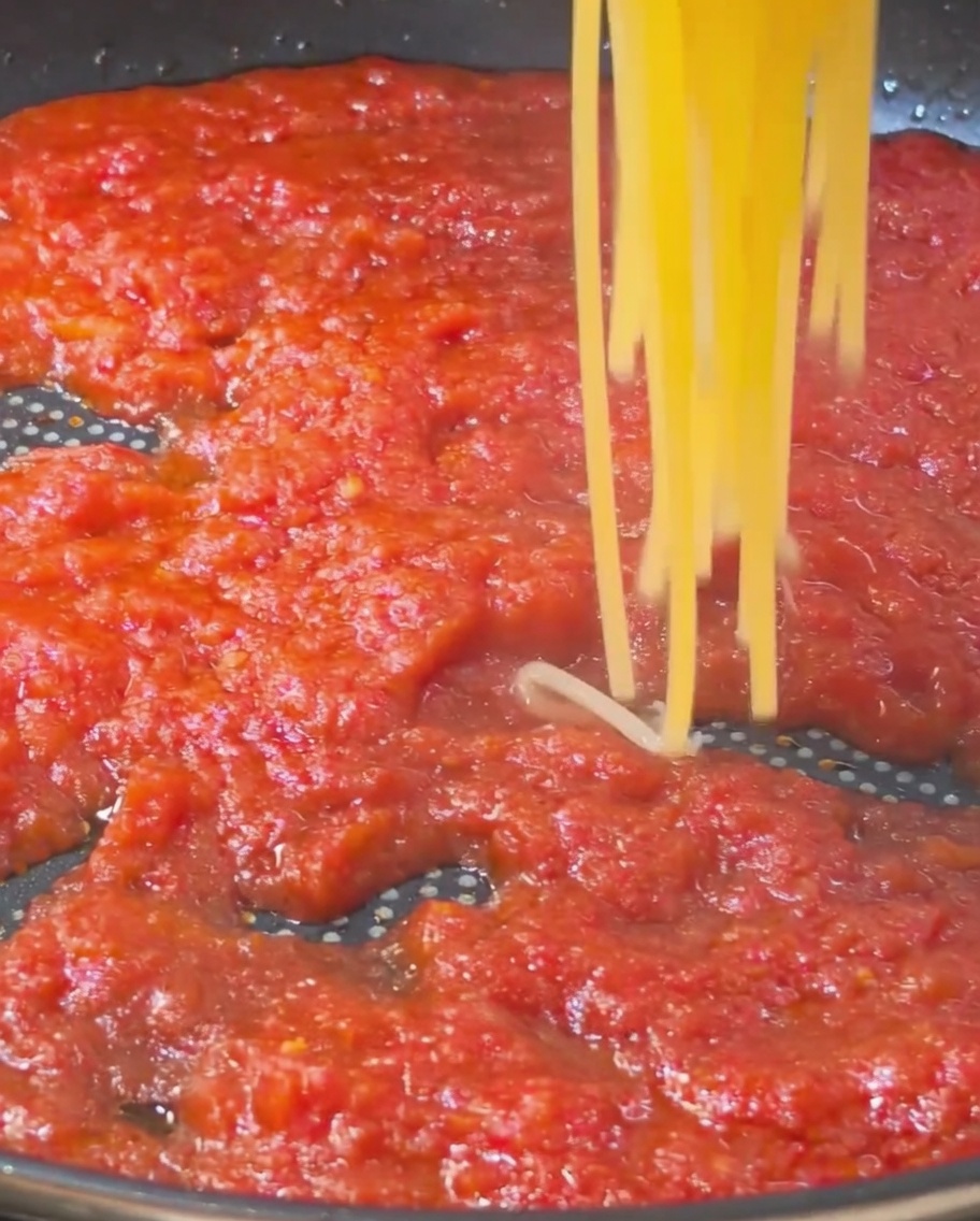 Spaghetti cotti e dorati sollevati e trasferiti in una padella piena di ricca salsa al pomodoro rossa.