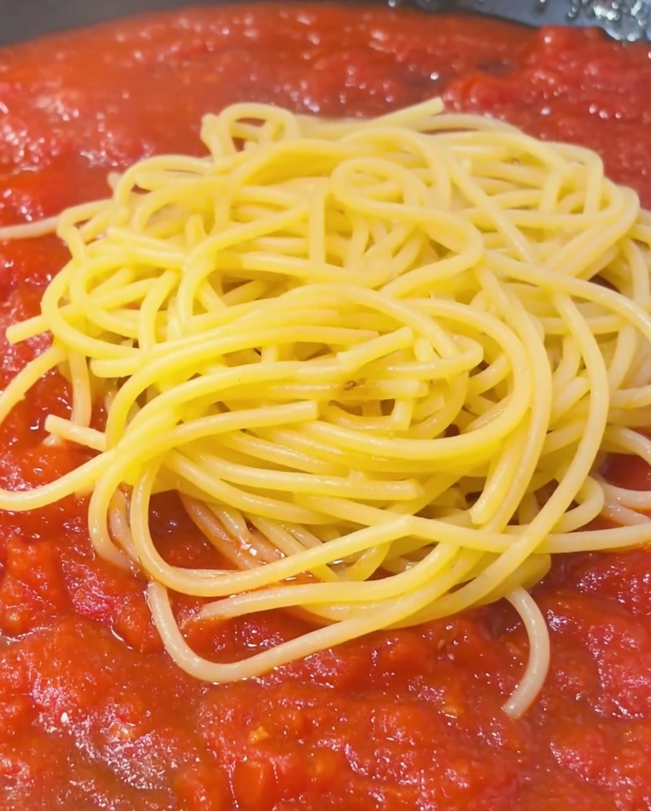 Un cucchiaio di acqua di cottura amidacea versato sopra un mucchio di spaghetti con salsa al pomodoro in padella.