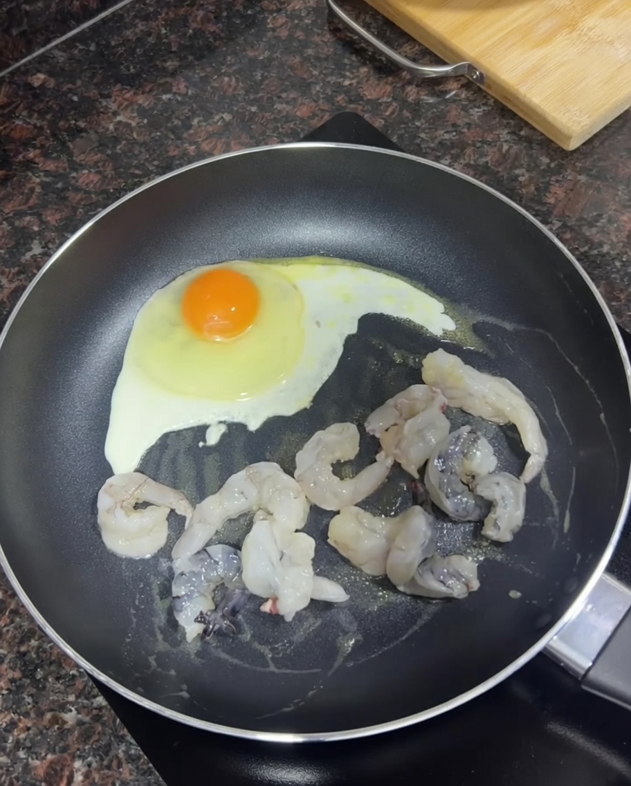 Gamberi crudi che dorano in padella accanto a un uovo che frigge.
